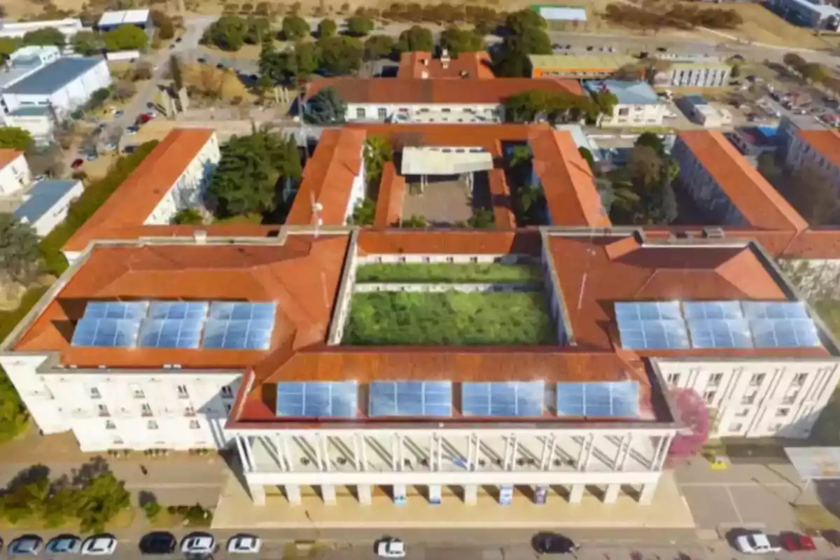 Aerial view of a large building with a red roof, solar panels, and a central courtyard with vegetation, surrounded by trees and cars parked in front.