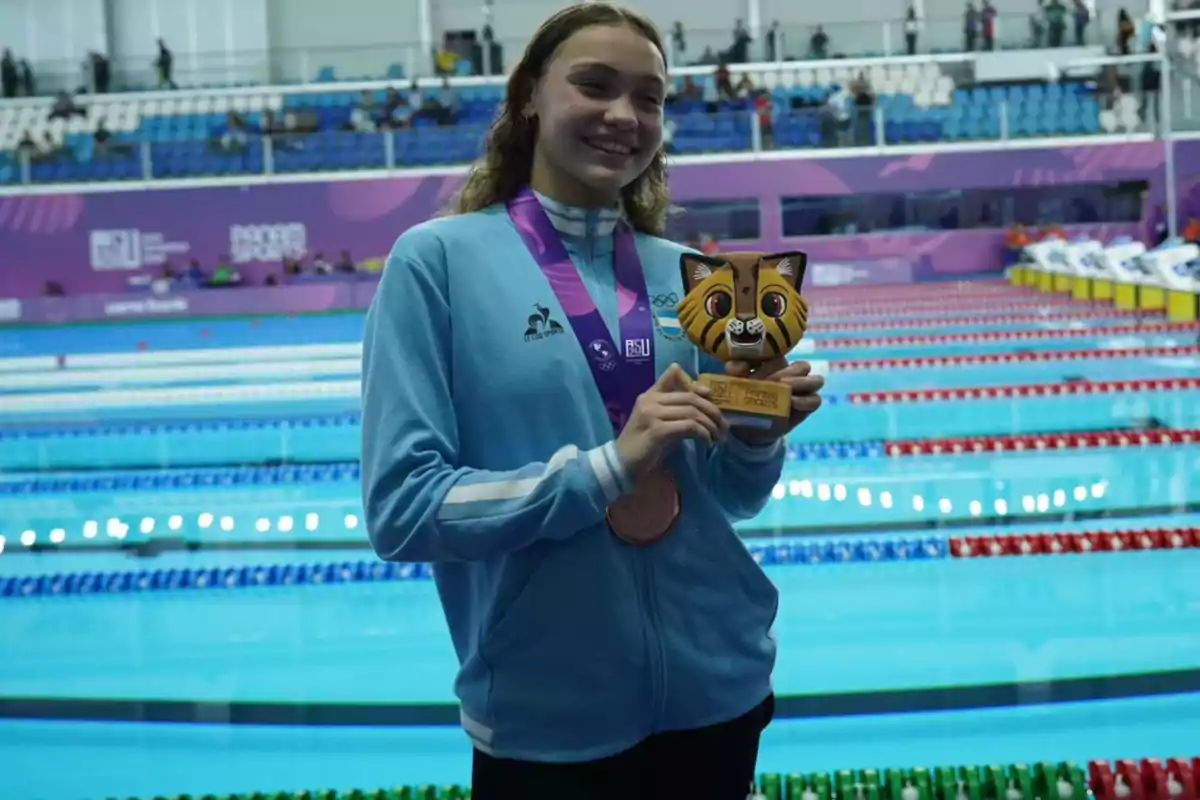 Joven nadadora sonriente sostiene un trofeo con forma de tigre y una medalla de bronce frente a una piscina olímpica Joven nadadora sonriente sostiene un trofeo con forma de tigre y una medalla de bronce frente a una piscina olímpica