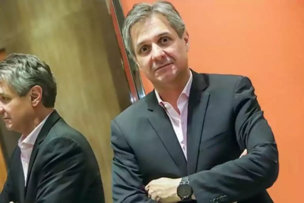 Gray-haired man wearing a dark jacket and light jersey posing in front of an orange wall and a mirror that reflects his image Gray-haired man wearing a dark jacket and light jersey posing in front of an orange wall and a mirror that reflects his image