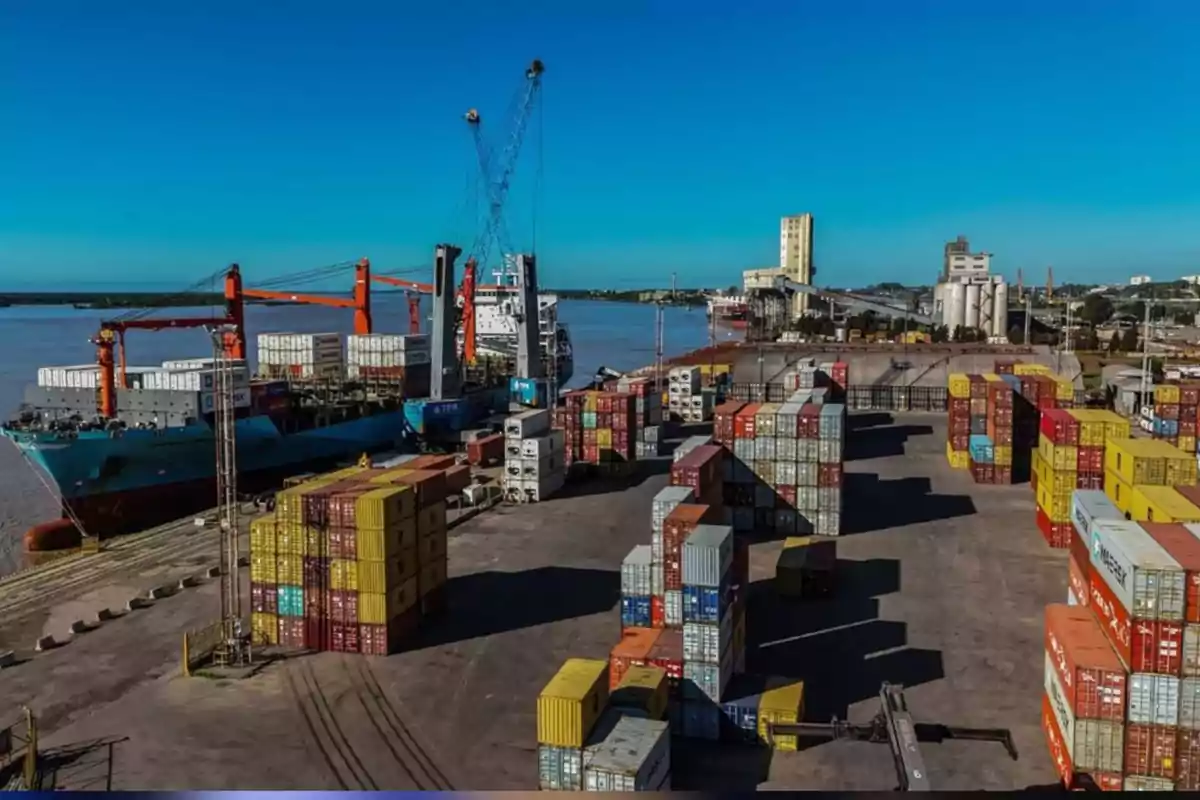 View of a commercial port with cranes, stacked containers, and cargo ships by the river under a clear sky