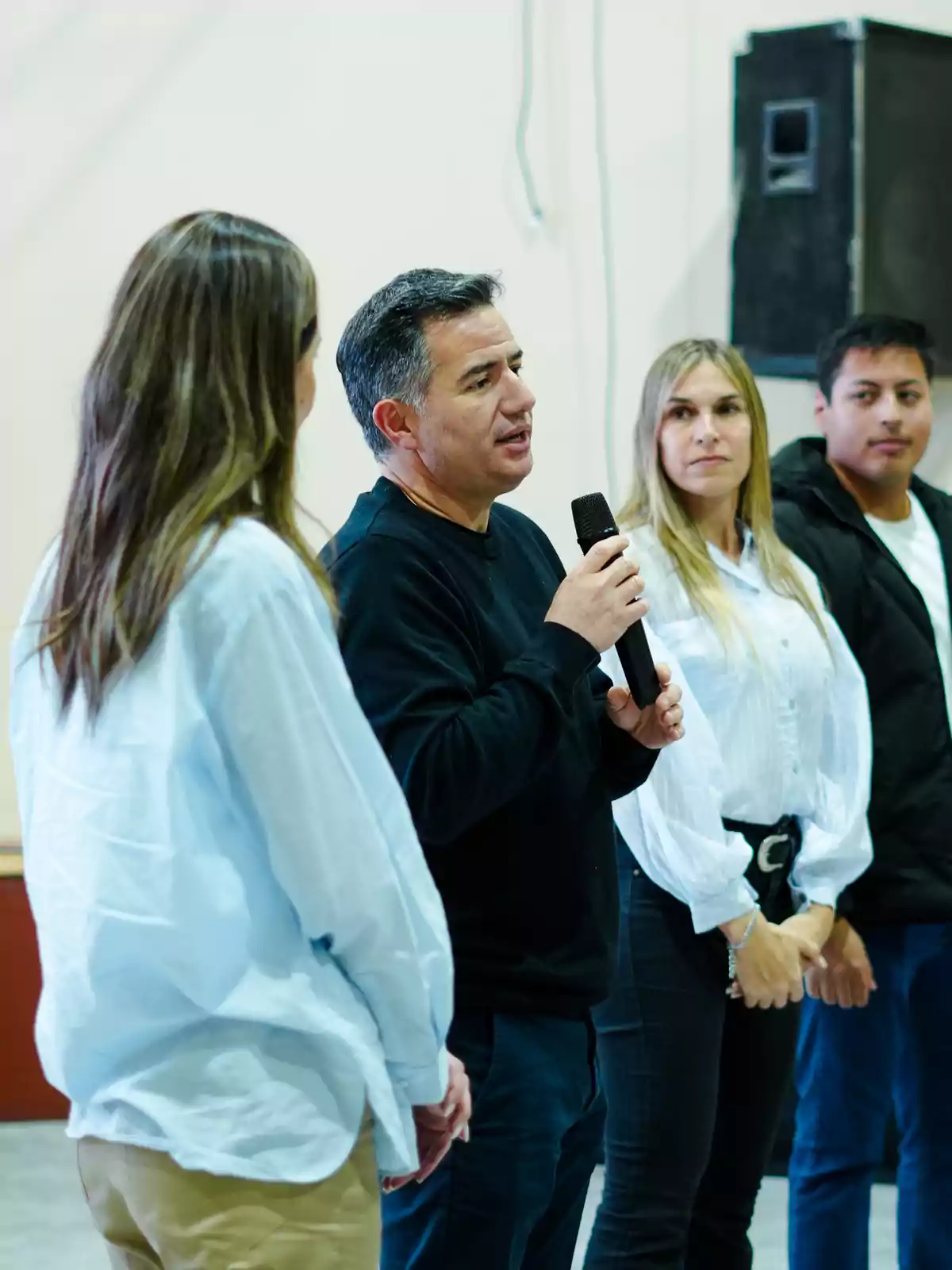 Un hombre sostiene un micrófono y habla mientras tres personas lo escuchan atentamente en un ambiente interior Un hombre sostiene un micrófono y habla mientras tres personas lo escuchan atentamente en un ambiente interior