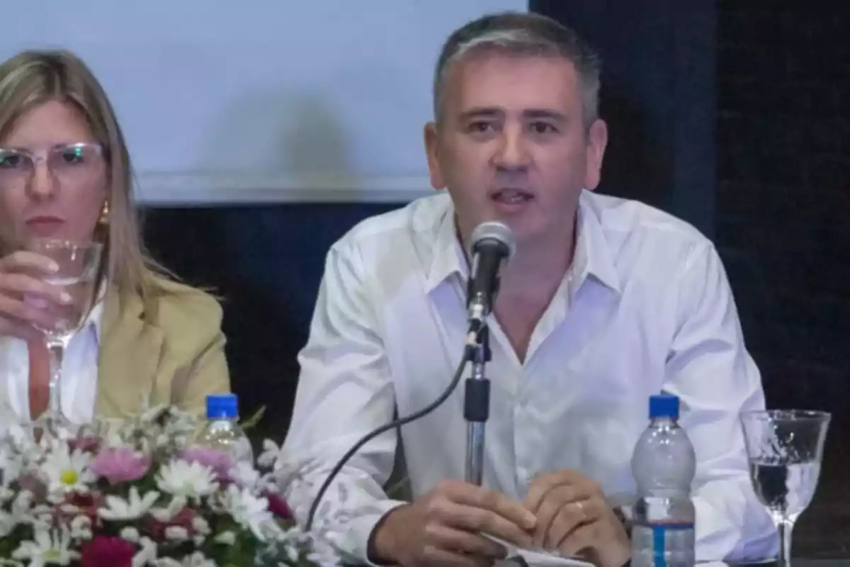 Hombre hablando en un micrófono durante una conferencia junto a una mujer que sostiene un vaso de agua, con flores y botellas de agua sobre la mesa