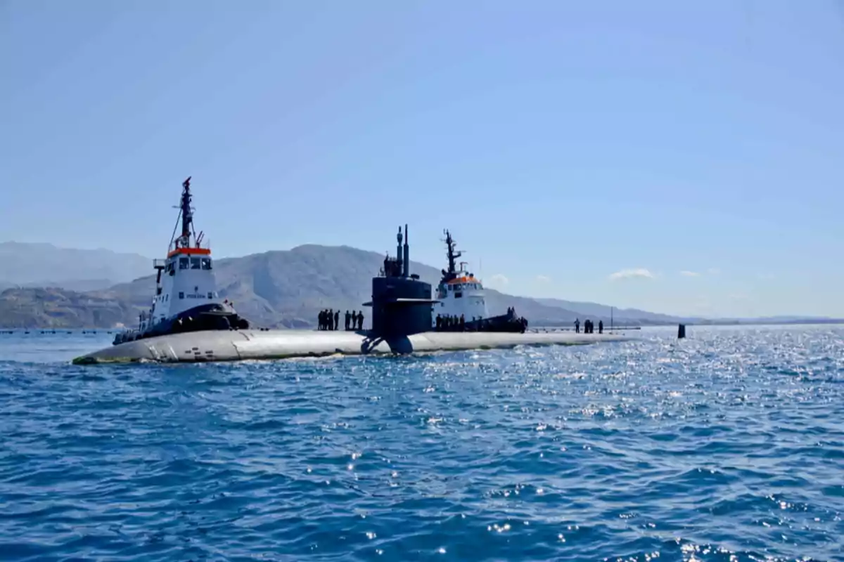 Submarino en la superficie del mar acompañado por dos barcos remolcadores con montañas al fondo y varias personas sobre el submarino Submarino en la superficie del mar acompañado por dos barcos remolcadores con montañas al fondo y varias personas sobre el submarino