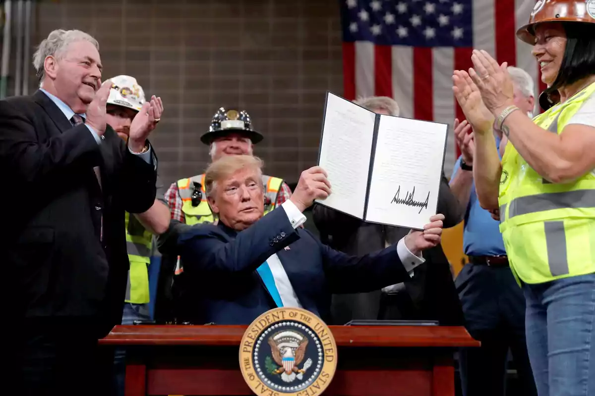 A man in a suit holds a signed document while several people around him, some wearing helmets and safety vests, applaud in front of a United States flag.