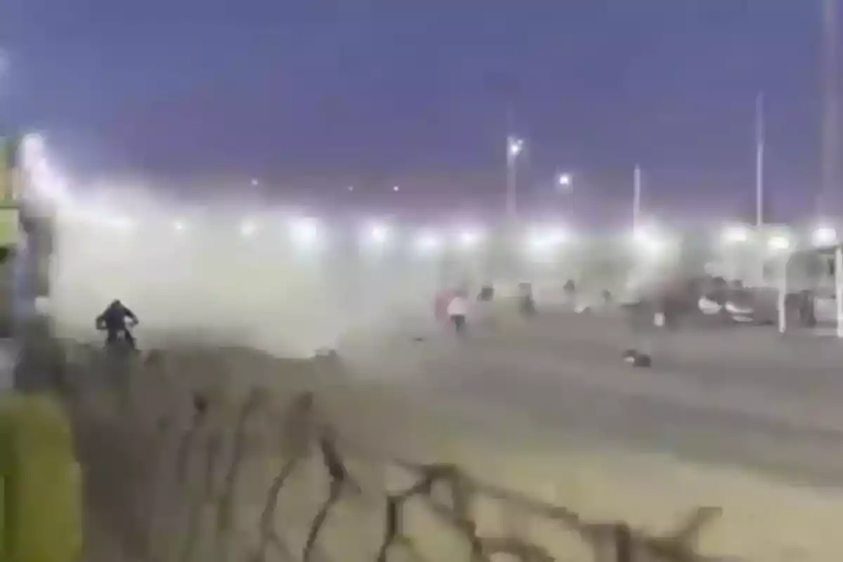 A motorcycle moves along a dusty track at sunset while several people watch from a distance and lights illuminate the background.