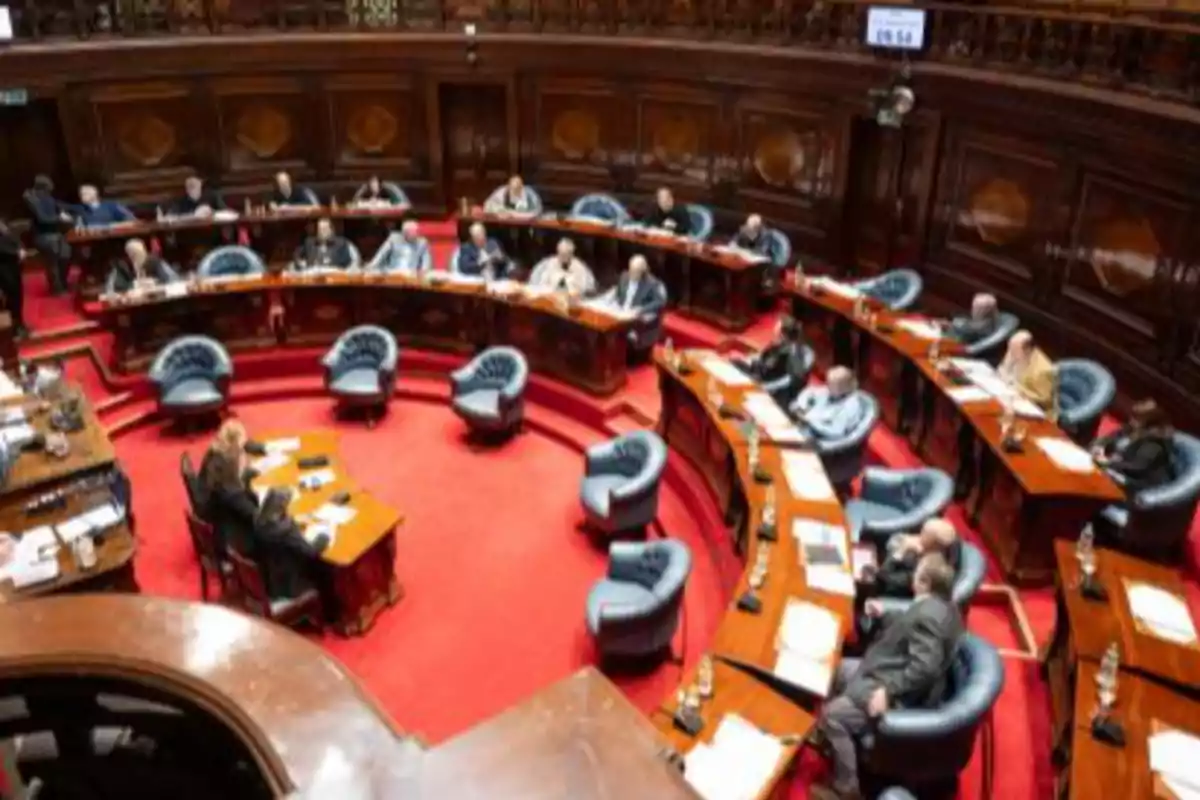 People seated in a parliamentary chamber with red carpet and wooden desks