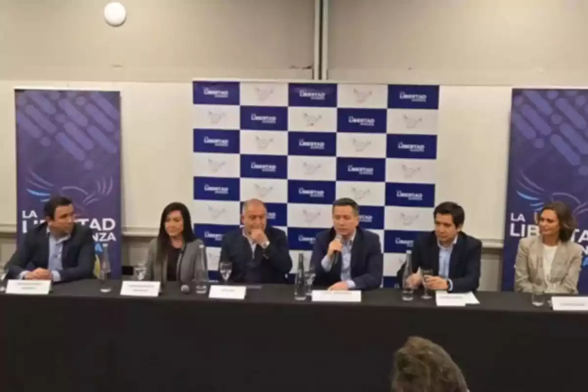 Six people sitting at a table during a press conference with a background of logos and banners that say La Libertad Avanza