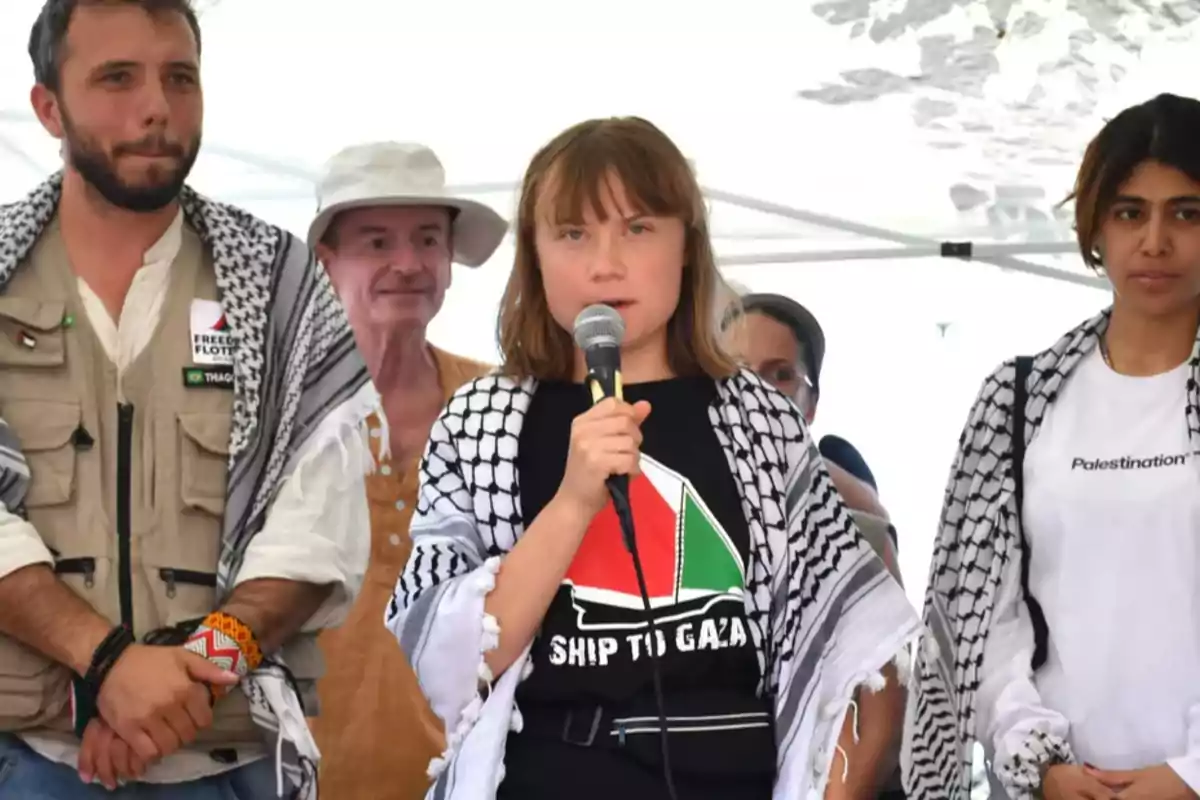 Una joven con micrófono y camiseta con el texto Ship to Gaza está acompañada por otras personas que llevan pañuelos palestinos en un evento público.