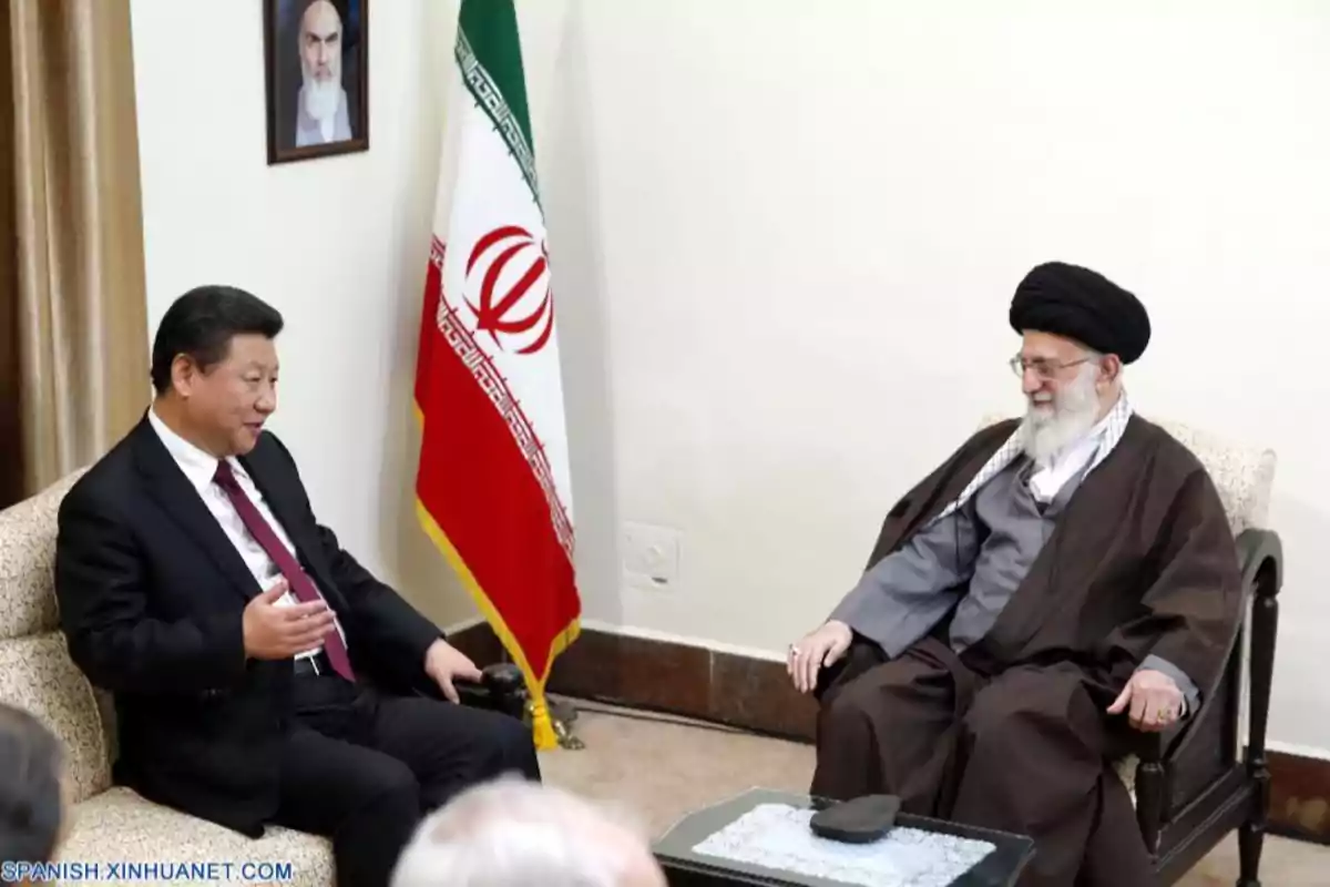 Dos hombres sentados conversando en una sala, con una bandera de Irán y un retrato en la pared al fondo