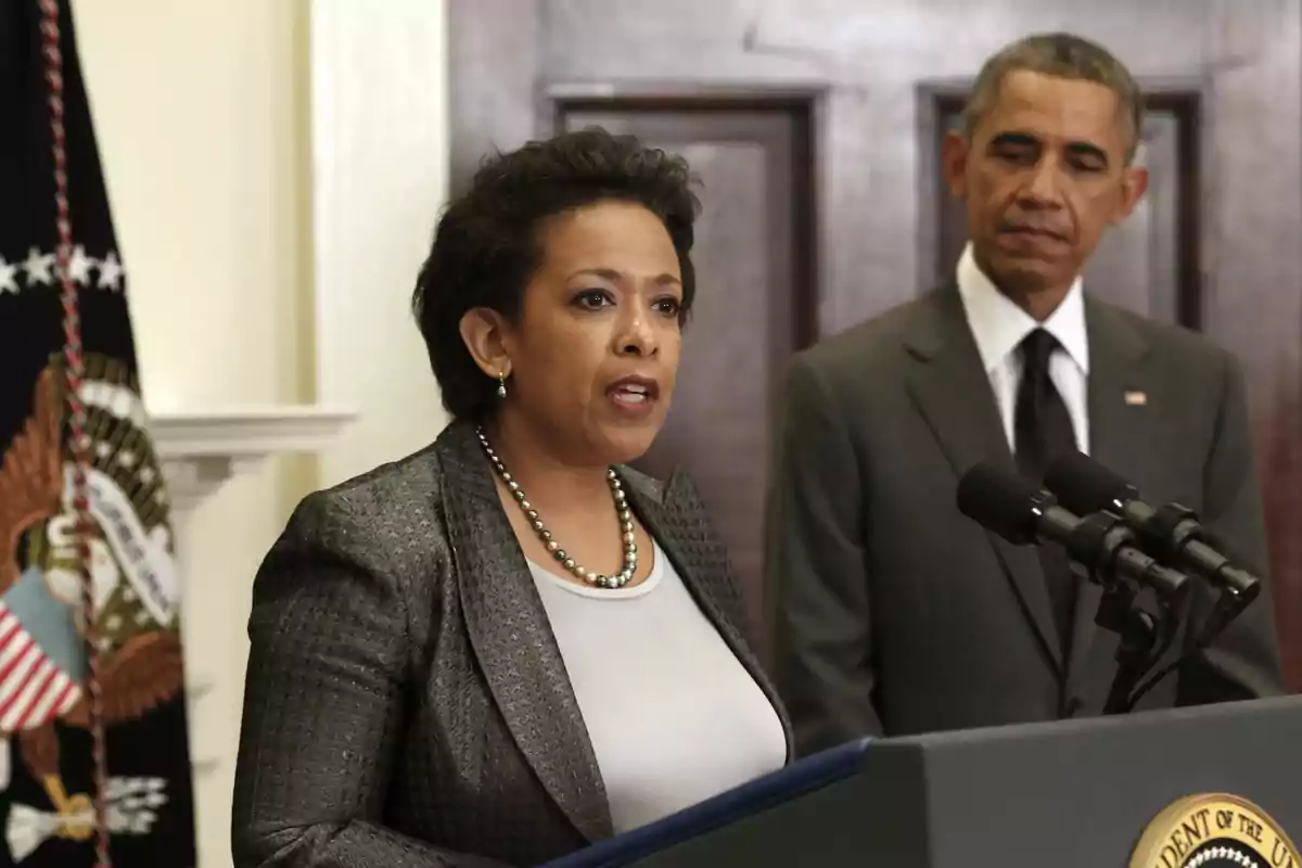 A woman speaks at a podium with microphones while a man in a suit watches her from the background.