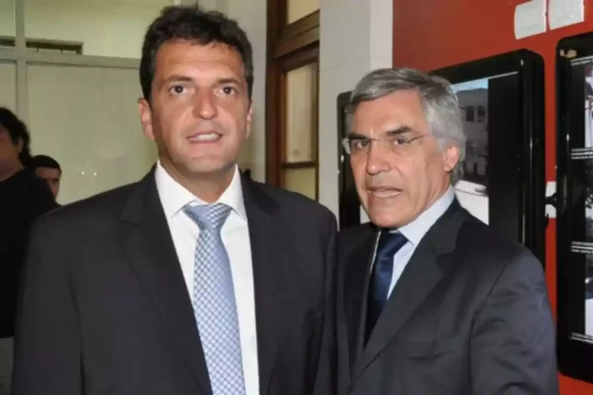 Two men dressed in suits and ties pose together in an indoor setting with red and white walls.