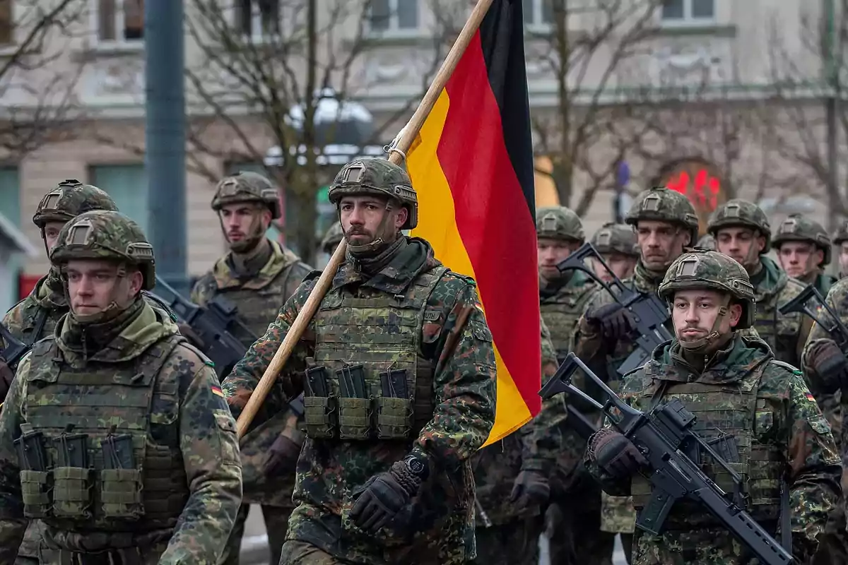 Soldados alemanes marchando en formación con uniforme de camuflaje y equipo militar, uno de ellos porta la bandera de Alemania