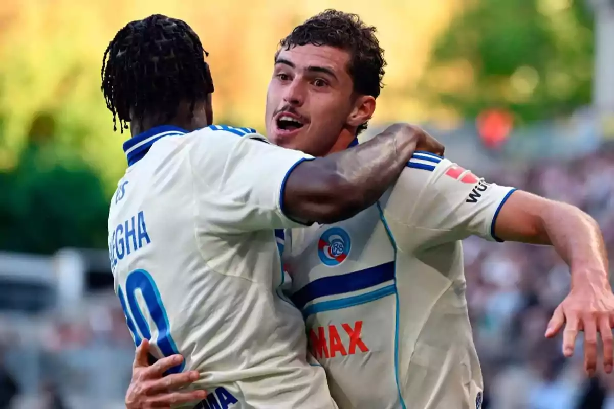 Two players in white uniforms celebrate a goal by hugging on the field. Two players in white uniforms celebrate a goal by hugging on the field.