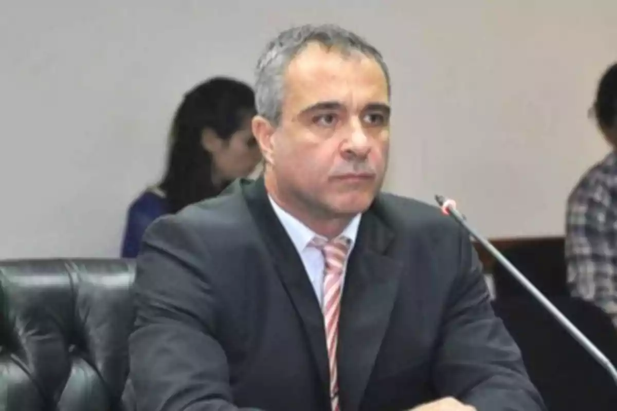 Man in a dark suit and tie sitting at a table in front of a microphone in a formal setting