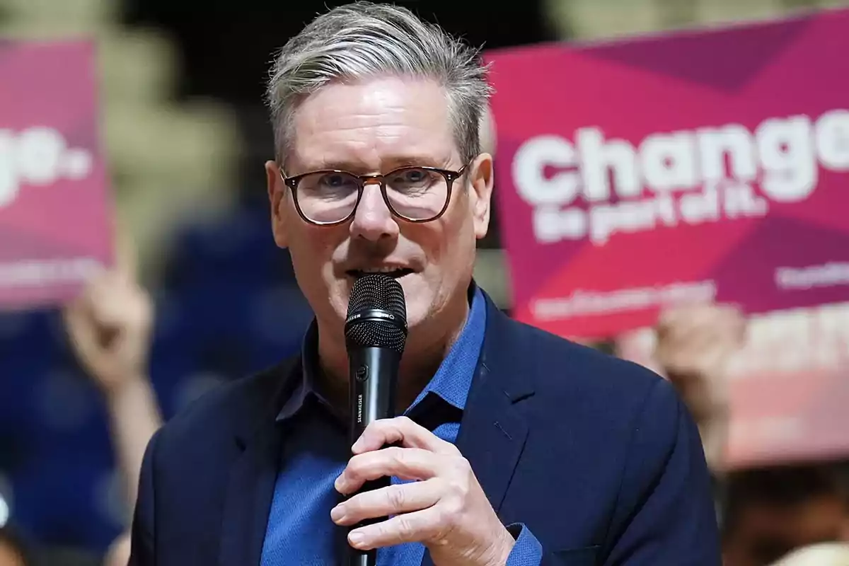 Gray-haired man with glasses speaking into a microphone at an event with signs in the background that say Change Gray-haired man with glasses speaking into a microphone at an event with signs in the background that say Change