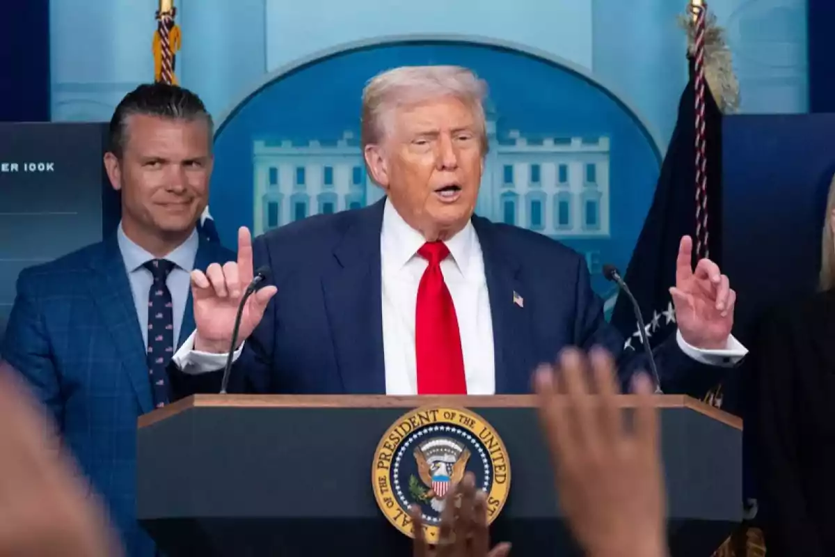 Un hombre de cabello rubio y corbata roja habla en un podio presidencial mientras varias manos se levantan frente a él y otras personas lo acompañan en el escenario