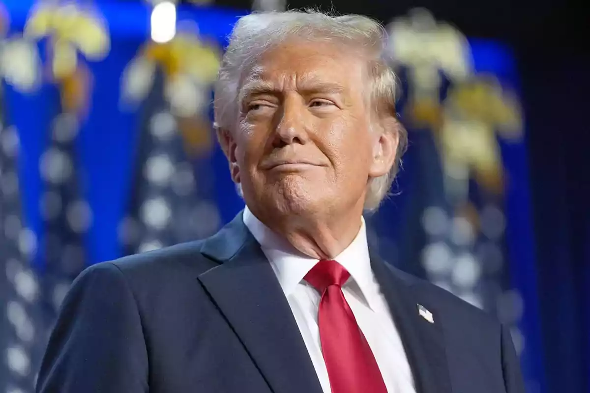 Blond-haired man in a dark suit and red tie in front of a background with blurred flags