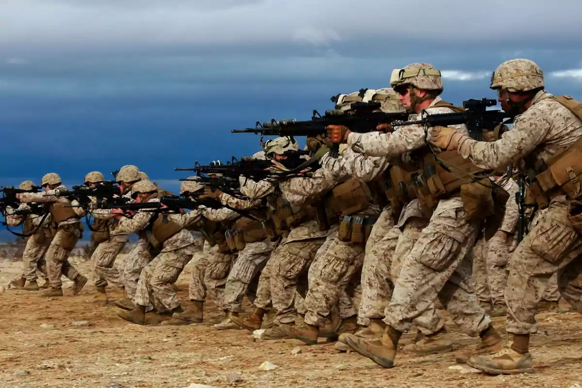 Soldados con uniforme de camuflaje apuntando sus rifles en formación sobre un terreno árido bajo un cielo nublado
