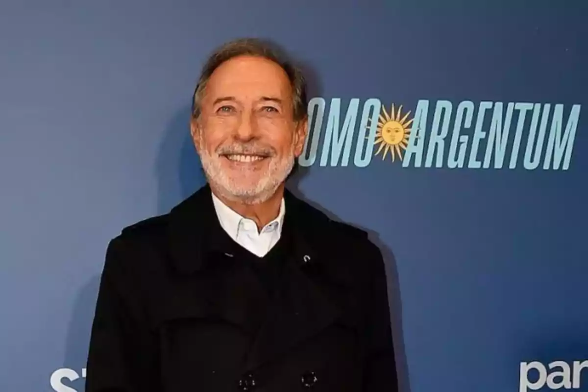 Gray-haired man with a smiling beard posing in front of a blue background with letters that say COMO ARGENTUM