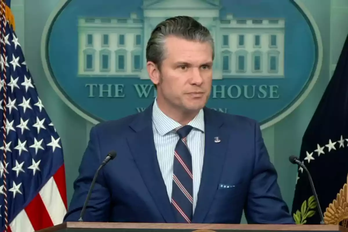 Man in a blue suit speaking at a podium in front of a backdrop with the White House logo and American flags