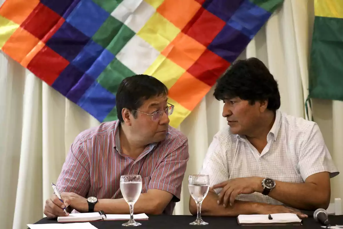 Two men are sitting at a table talking, with glasses of water and notebooks; in the background, there is a multicolored flag and another green and yellow flag. Two men are sitting at a table talking, with glasses of water and notebooks; in the background, there is a multicolored flag and another green and yellow flag.