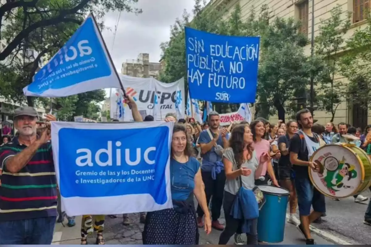 People marching down the street with signs and flags in defense of public education, some people playing drums, and a peaceful protest atmosphere can be observed. People marching down the street with signs and flags in defense of public education, some people playing drums, and a peaceful protest atmosphere can be observed.