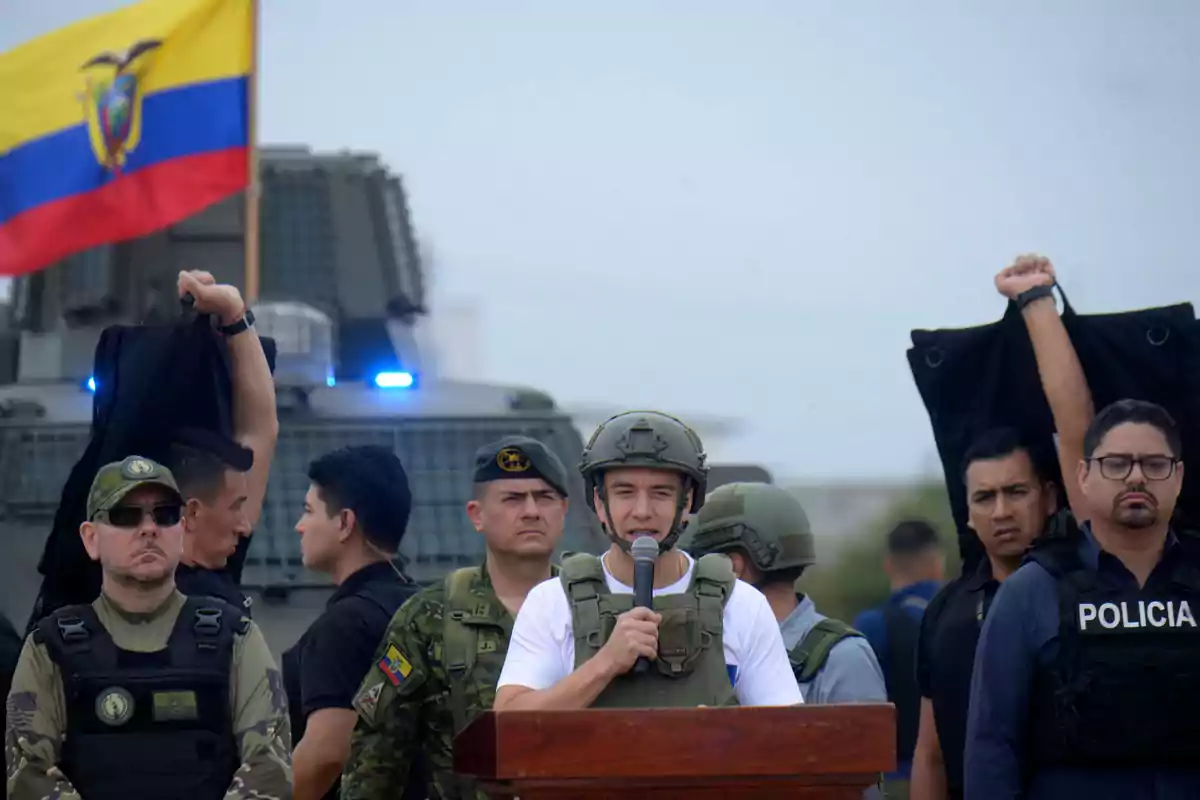 Un grupo de militares y policías rodea a un hombre con casco y chaleco antibalas que habla frente a un micrófono mientras al fondo ondea una bandera de Ecuador