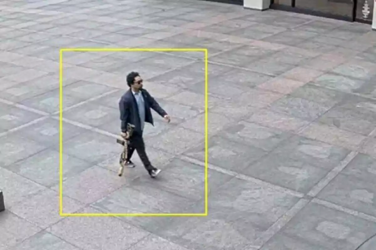 Man walking in a concrete plaza holding a large rifle-shaped object inside a yellow box Man walking in a concrete plaza holding a large rifle-shaped object inside a yellow box
