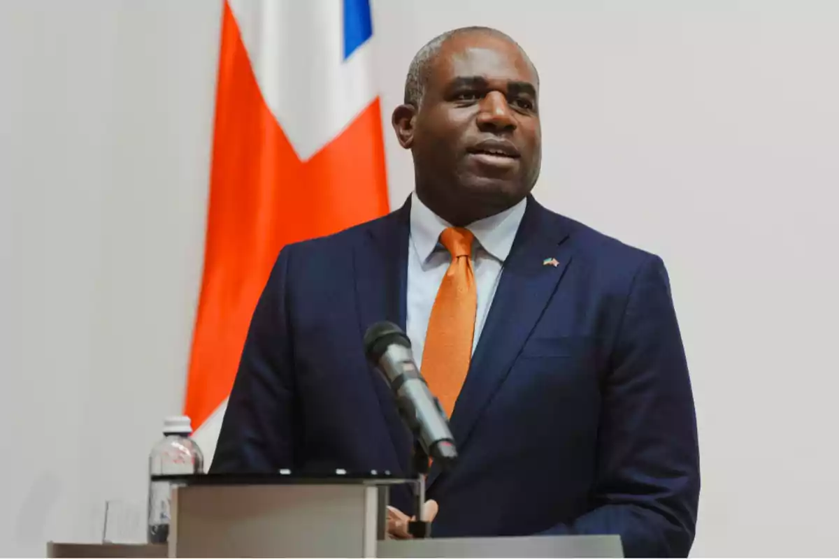 Hombre de traje azul y corbata naranja hablando frente a un micrófono con una bandera de fondo Hombre de traje azul y corbata naranja hablando frente a un micrófono con una bandera de fondo