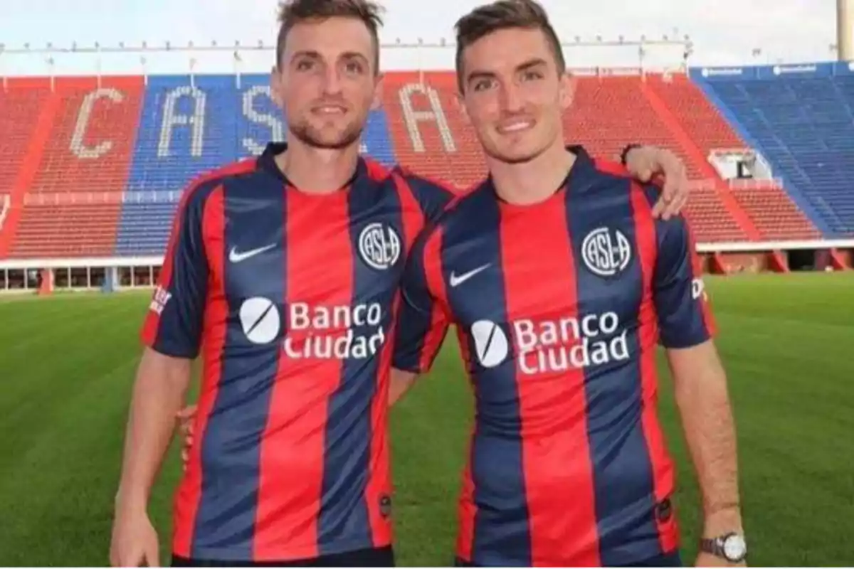 Dos futbolistas con uniforme de San Lorenzo posan abrazados en el estadio con gradas rojas y azules de fondo Dos futbolistas con uniforme de San Lorenzo posan abrazados en el estadio con gradas rojas y azules de fondo