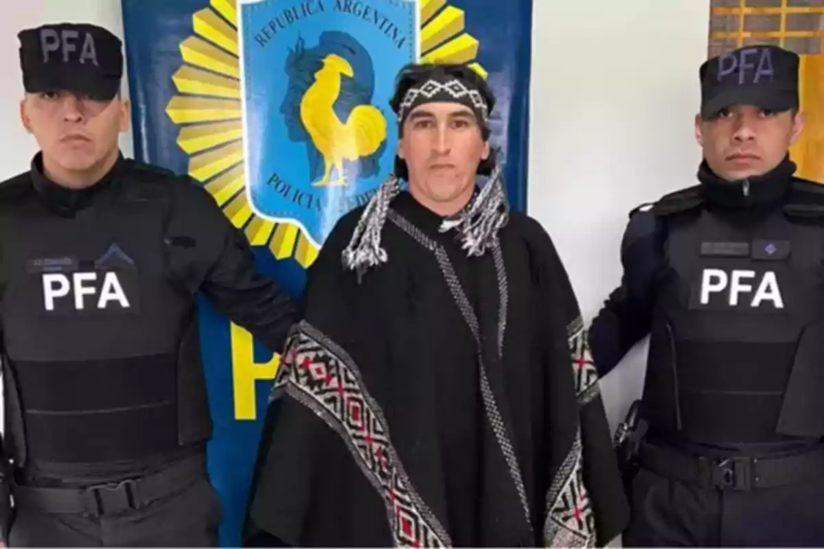 Tres personas posan frente a un cartel de la Policía Federal Argentina, dos de ellas con uniforme policial y la persona del centro con vestimenta tradicional mapuche.