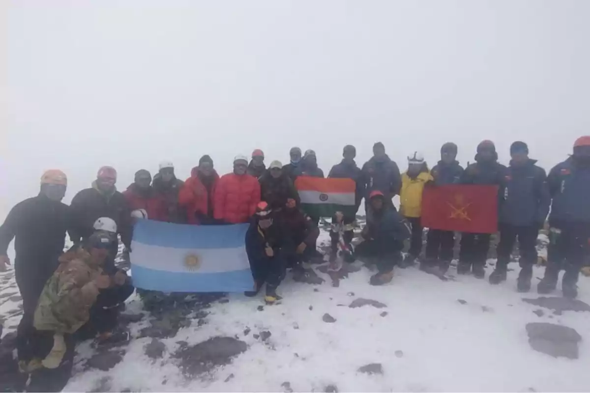 Grupo de montañistas posando en la cima nevada de una montaña con banderas de Argentina, India y una bandera roja