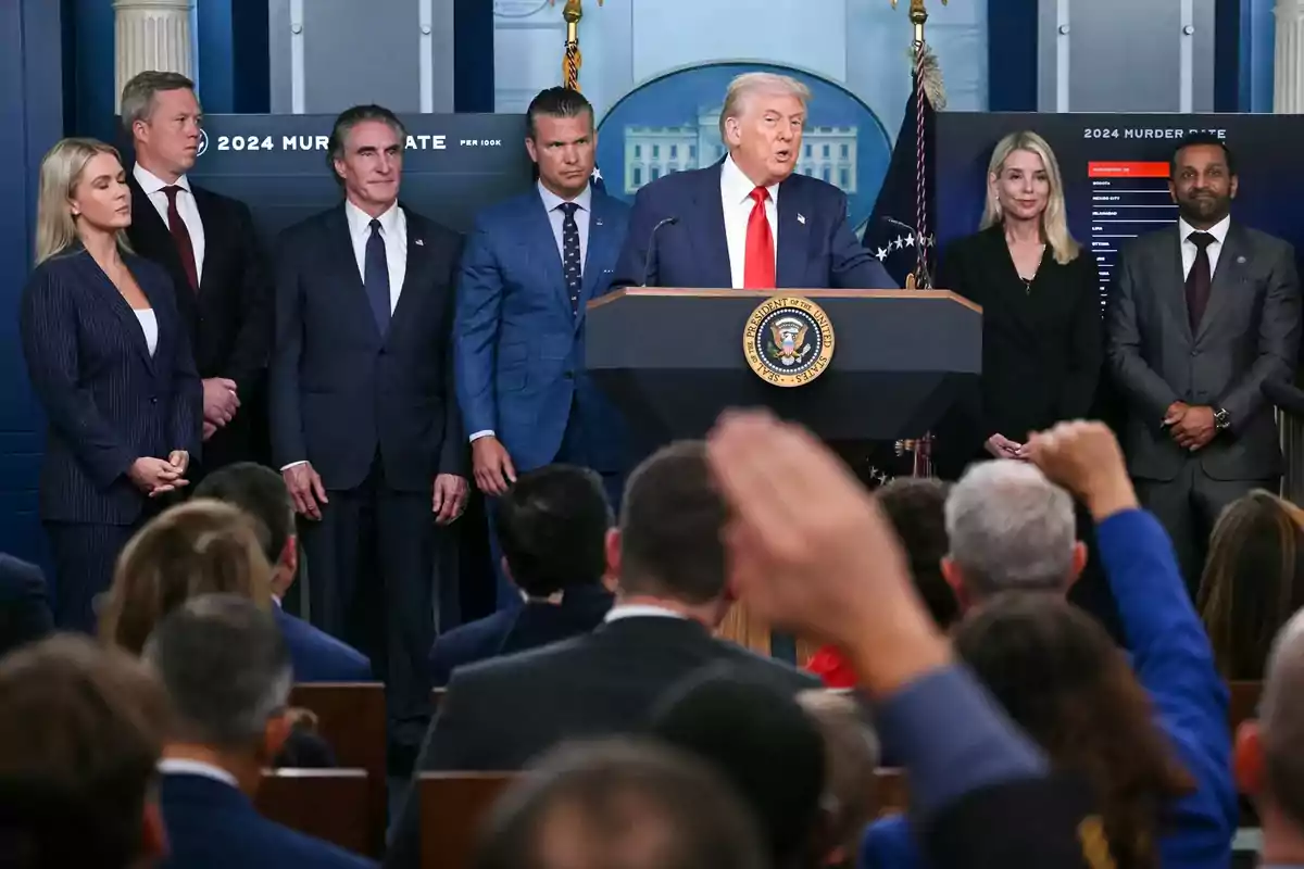 A group of people standing behind a presidential podium while a blond-haired man speaks to an audience in a conference room.