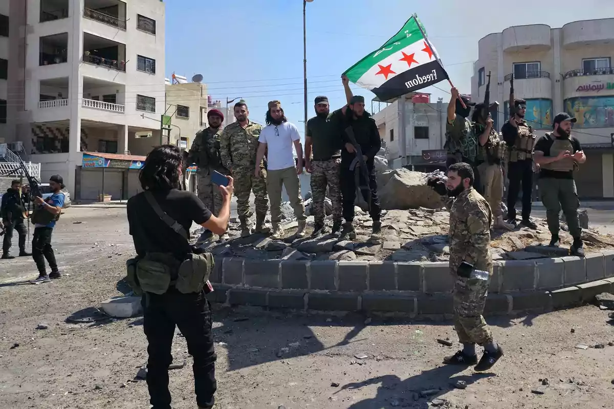 A group of armed men wearing military and civilian clothing pose on a stone structure in an urban street while one of them holds a flag and another takes a photo with his cellphone.
