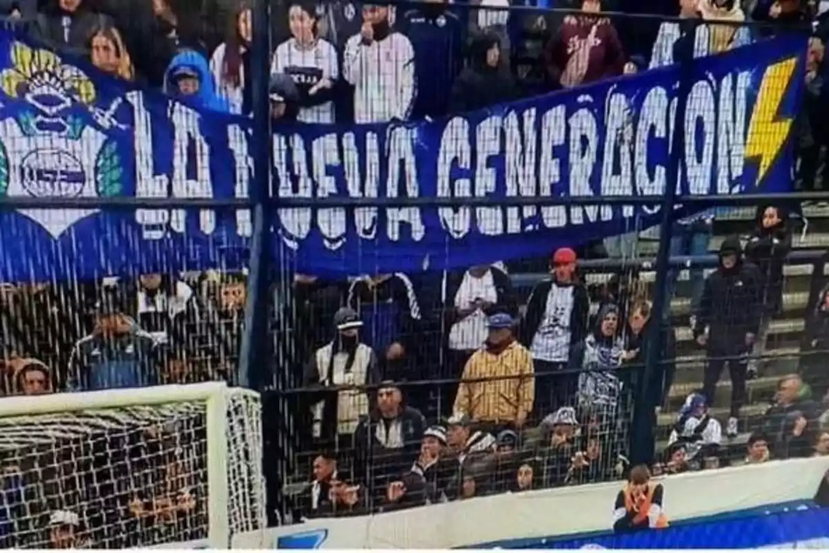 Aficionados en una tribuna de estadio con una bandera azul que dice La Nueva Generación y un rayo amarillo Aficionados en una tribuna de estadio con una bandera azul que dice La Nueva Generación y un rayo amarillo