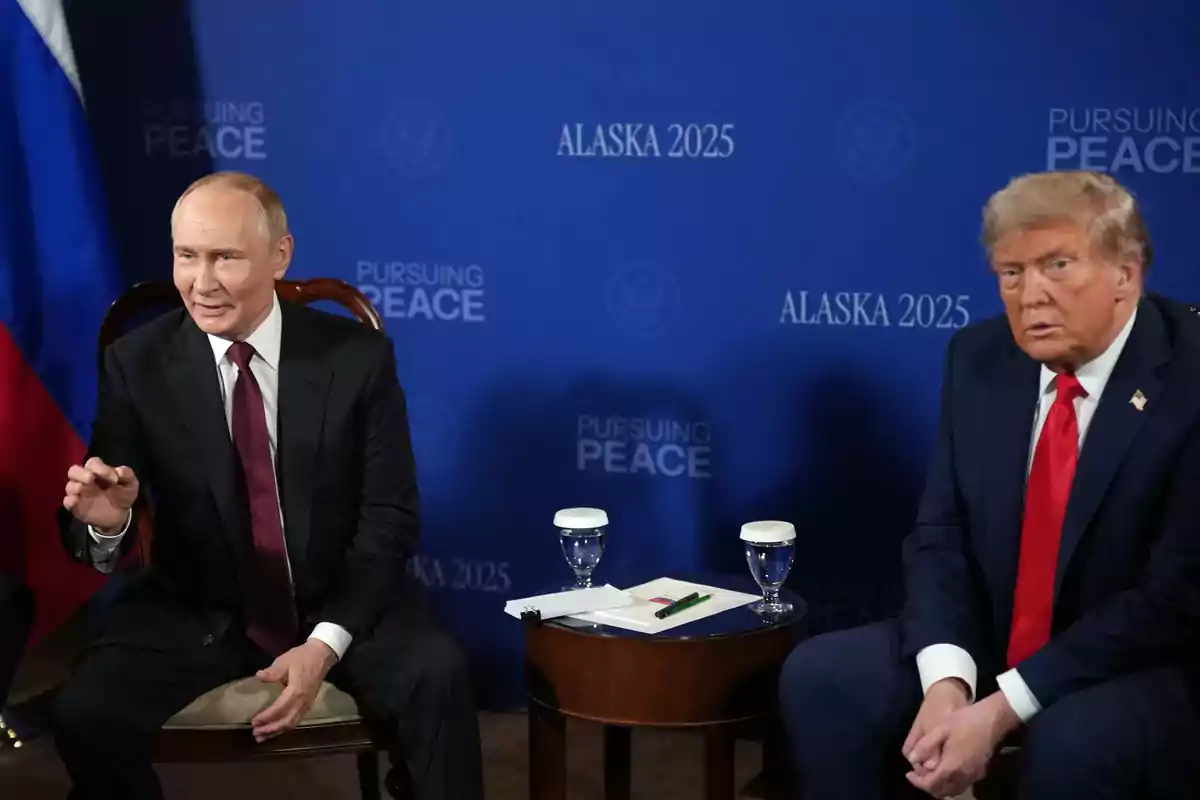 Two men in dark suits and ties sitting next to a small table with two glasses of water and papers, in front of a blue background that says Alaska 2025 and Pursuing Peace. Two men in dark suits and ties sitting next to a small table with two glasses of water and papers, in front of a blue background that says Alaska 2025 and Pursuing Peace.