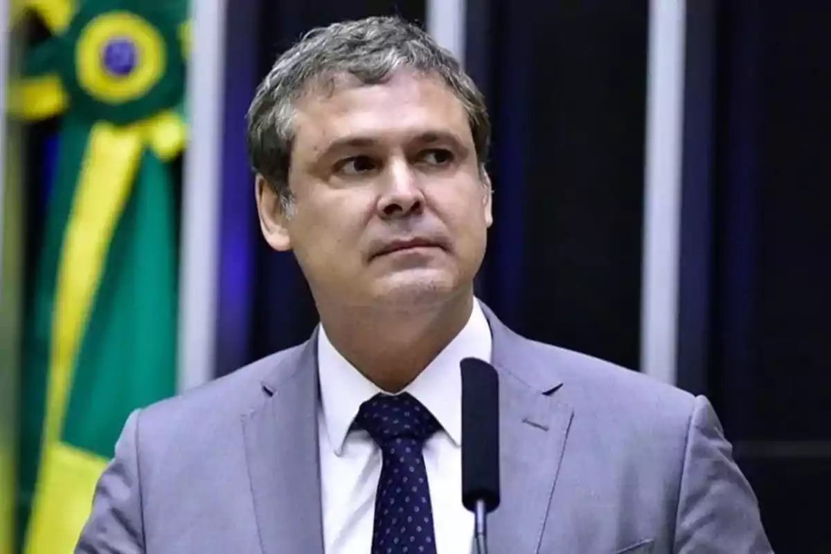 Short-haired, gray-haired man in a gray suit and blue polka-dot tie speaking in front of a microphone with a green and yellow flag in the background Short-haired, gray-haired man in a gray suit and blue polka-dot tie speaking in front of a microphone with a green and yellow flag in the background