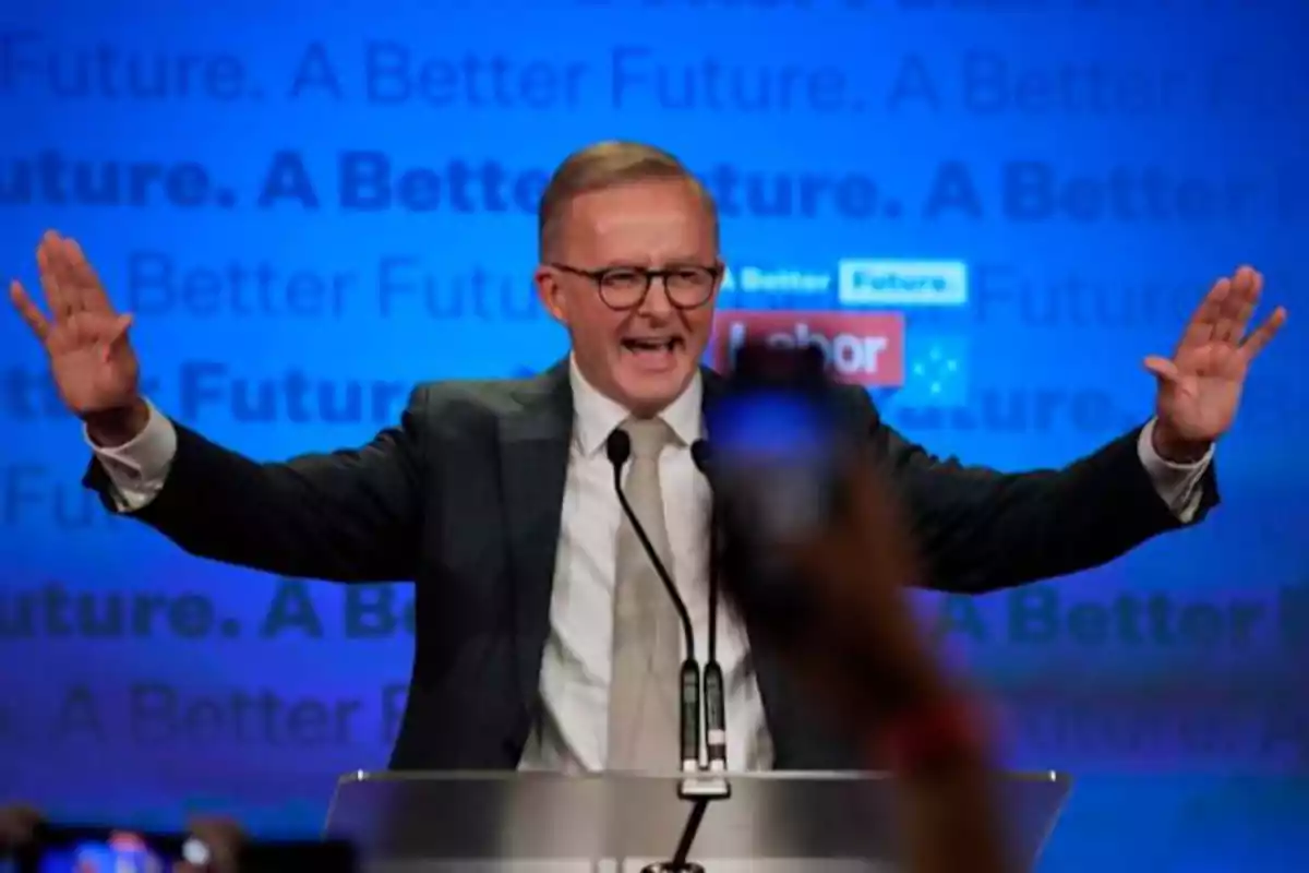 Man with glasses and suit speaking at a podium with a blue background and the words Man with glasses and suit speaking at a podium with a blue background and the words