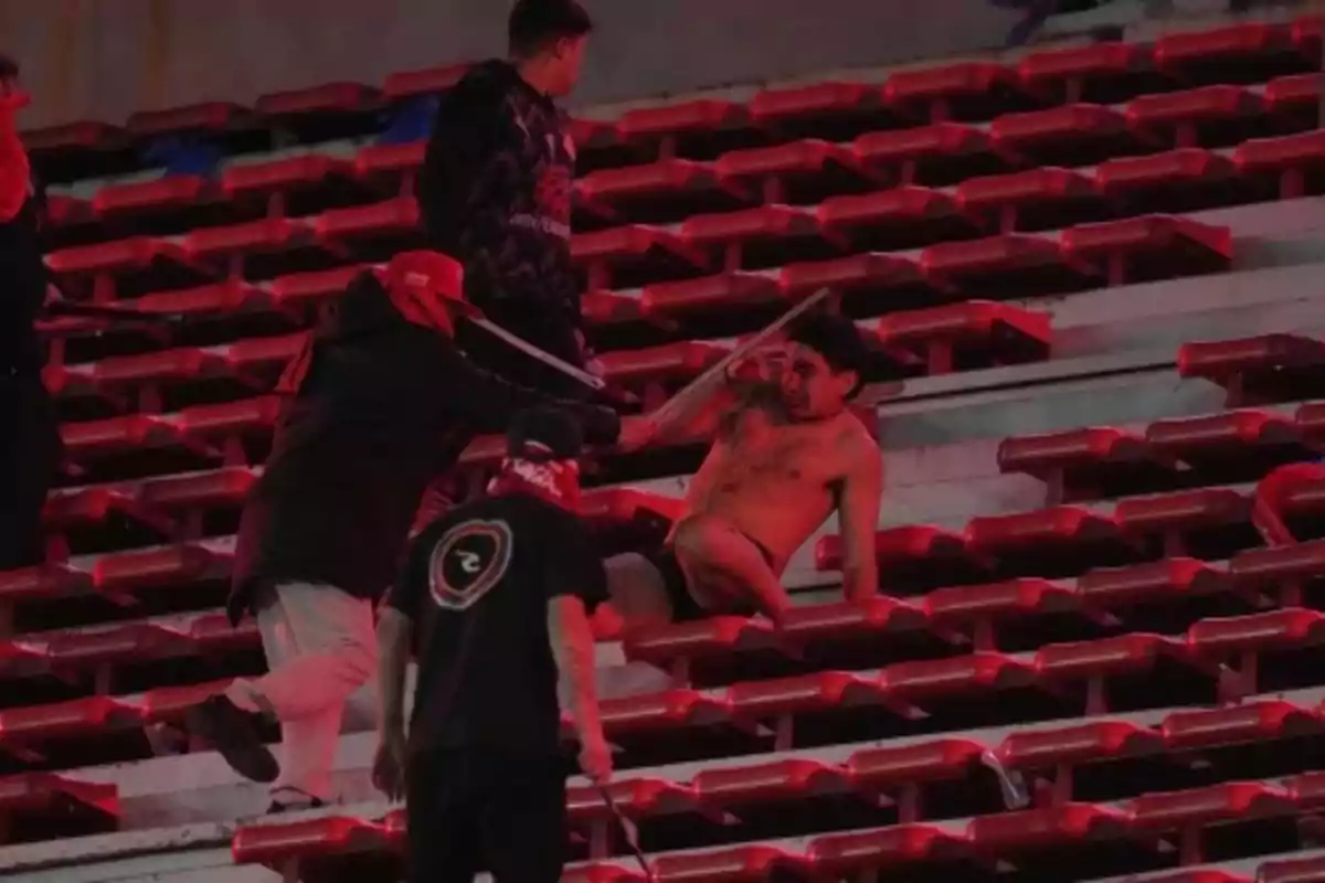 A jerseyless man is assaulted by several people in the stands of a stadium with red seats. A jerseyless man is assaulted by several people in the stands of a stadium with red seats.