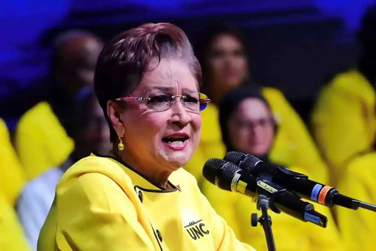 Older woman with glasses and yellow clothing speaking in front of several microphones at an event with people dressed in yellow in the background Older woman with glasses and yellow clothing speaking in front of several microphones at an event with people dressed in yellow in the background