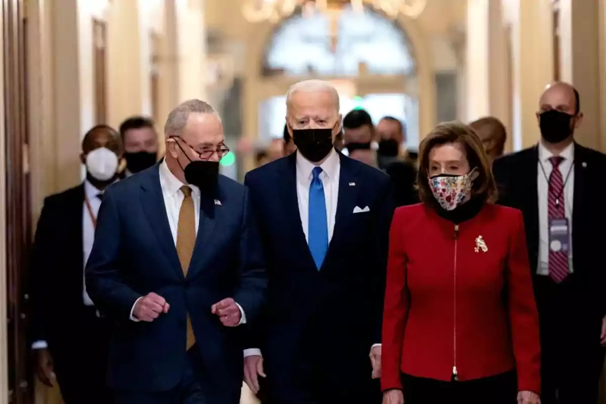 Three people wearing face masks walk down an elegant hallway accompanied by several others in the background.