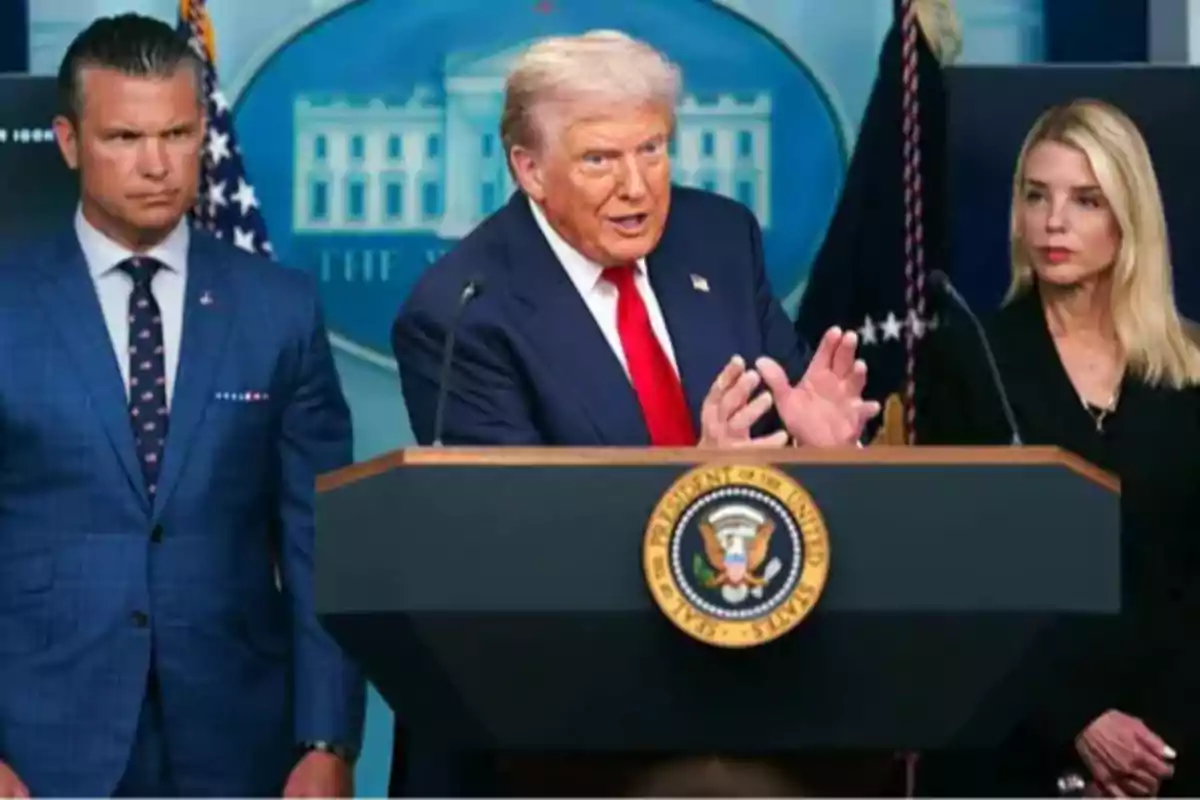 Tres personas de pie en una sala de prensa oficial con un podio presidencial al frente y el escudo de la Casa Blanca en el fondo Tres personas de pie en una sala de prensa oficial con un podio presidencial al frente y el escudo de la Casa Blanca en el fondo