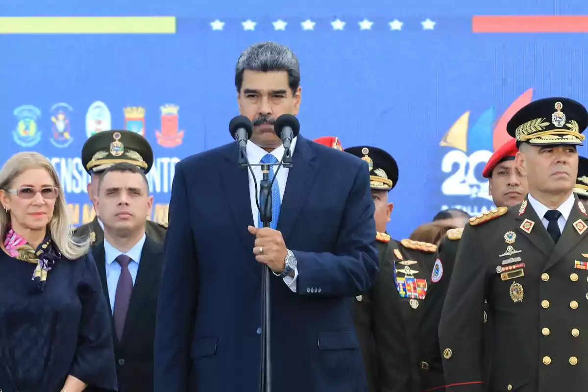 A man in a blue suit speaks in front of two microphones, surrounded by military personnel and a woman with glasses at an official event. A man in a blue suit speaks in front of two microphones, surrounded by military personnel and a woman with glasses at an official event.