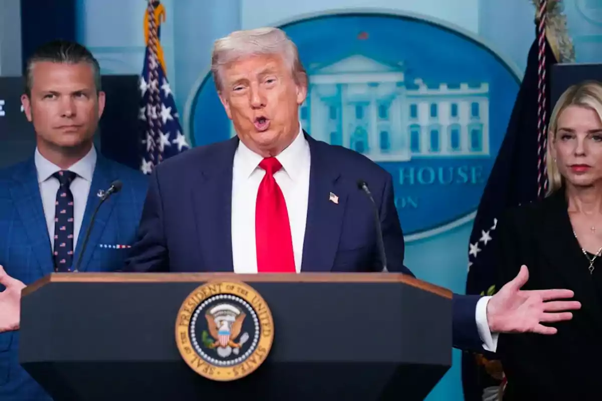 Hombre de cabello rubio con corbata roja hablando en un podio presidencial acompañado por dos personas en un escenario oficial