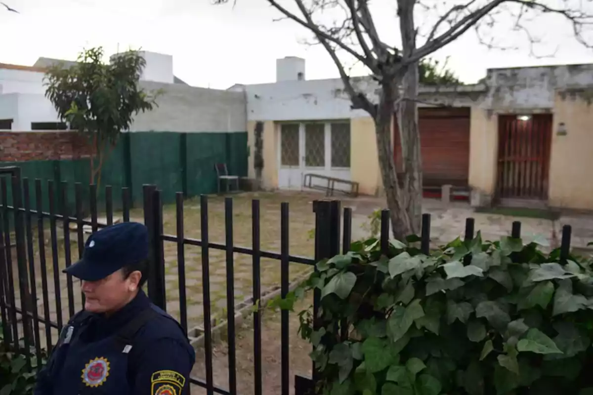 Una mujer policía con uniforme oscuro está de pie frente a una reja negra que rodea el patio de una casa de una sola planta con paredes claras y ventanas enrejadas, al fondo hay un árbol sin hojas y vegetación en primer plano