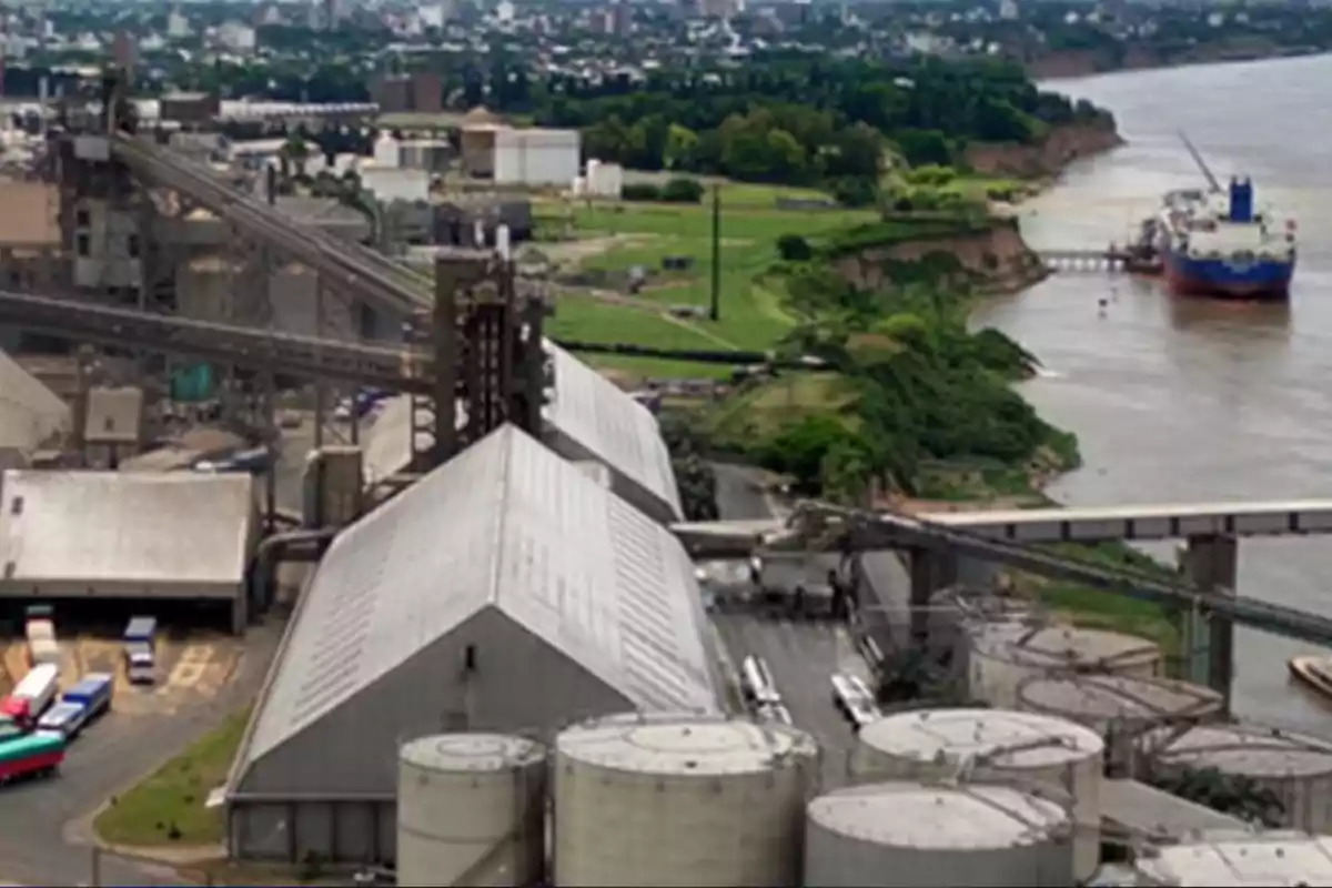 Vista aérea de un complejo industrial junto a un río con silos, depósitos, camiones y un barco atracado en el muelle
