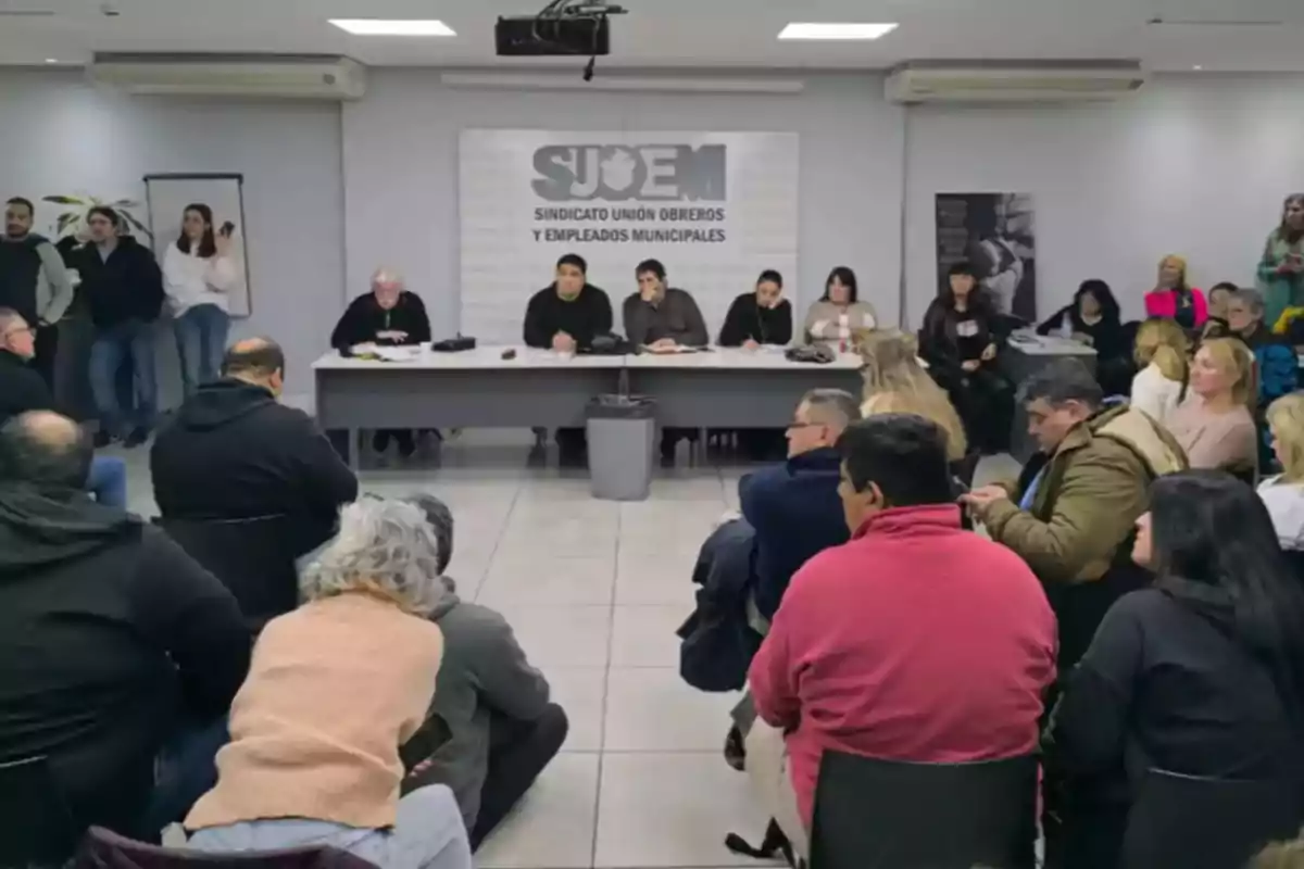 Reunión de personas en una sala con varias personas sentadas frente a una mesa larga y un cartel de sindicato en la pared Reunión de personas en una sala con varias personas sentadas frente a una mesa larga y un cartel de sindicato en la pared