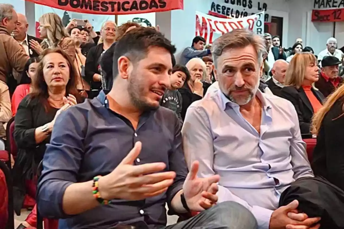 Two men are talking while seated in an auditorium full of people with signs and a gathering or protest atmosphere. Two men are talking while seated in an auditorium full of people with signs and a gathering or protest atmosphere.