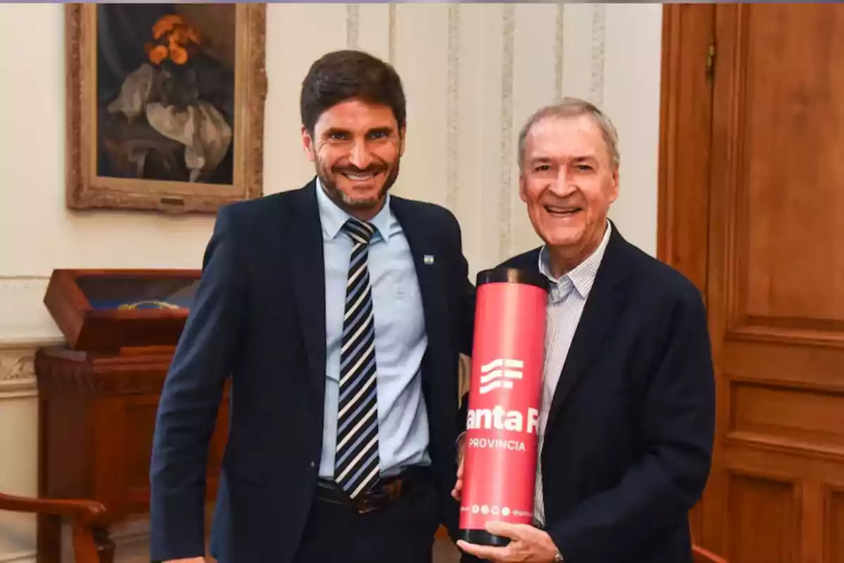 Dos hombres sonrientes posan en una oficina elegante mientras uno de ellos sostiene un termo rojo con el logo de la provincia de Santa Fe Dos hombres sonrientes posan en una oficina elegante mientras uno de ellos sostiene un termo rojo con el logo de la provincia de Santa Fe