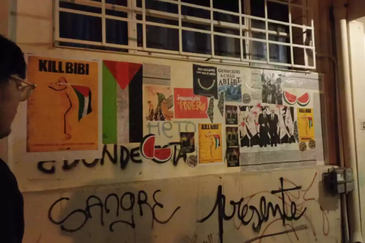 Un hombre observa una pared cubierta de carteles políticos, banderas y grafitis en una calle durante la noche Un hombre observa una pared cubierta de carteles políticos, banderas y grafitis en una calle durante la noche